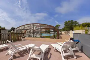 Terrasse eines Campingplatzes mit mehreren weißen Liegestühlen, Sonnenschutzdach und umzäuntem Bereich im Hintergrund.