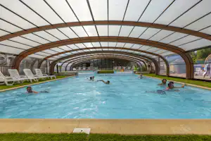 Schwimmbecken im Campingbereich unter einem Glasdach, mit Liegestühlen am Beckenrand und mehreren Personen im Wasser.