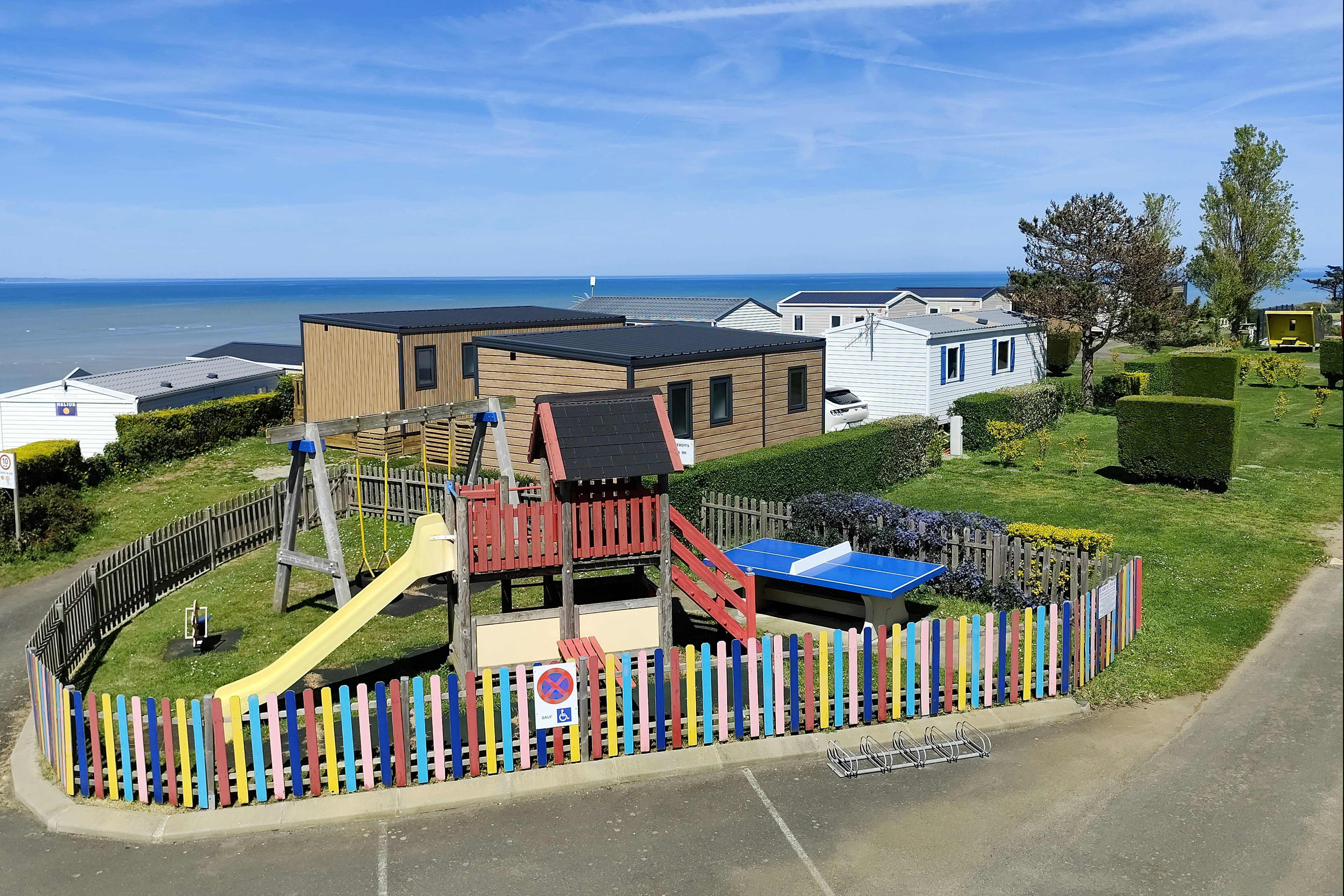 Spielplatz mit Rutsche und Klettergerüst vor Ferienhäusern, Blick auf das Meer und den Himmel über dem Campingplatz.