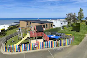 Spielplatz mit Rutsche und Klettergerüst vor Ferienhäusern, Blick auf das Meer und den Himmel über dem Campingplatz.
