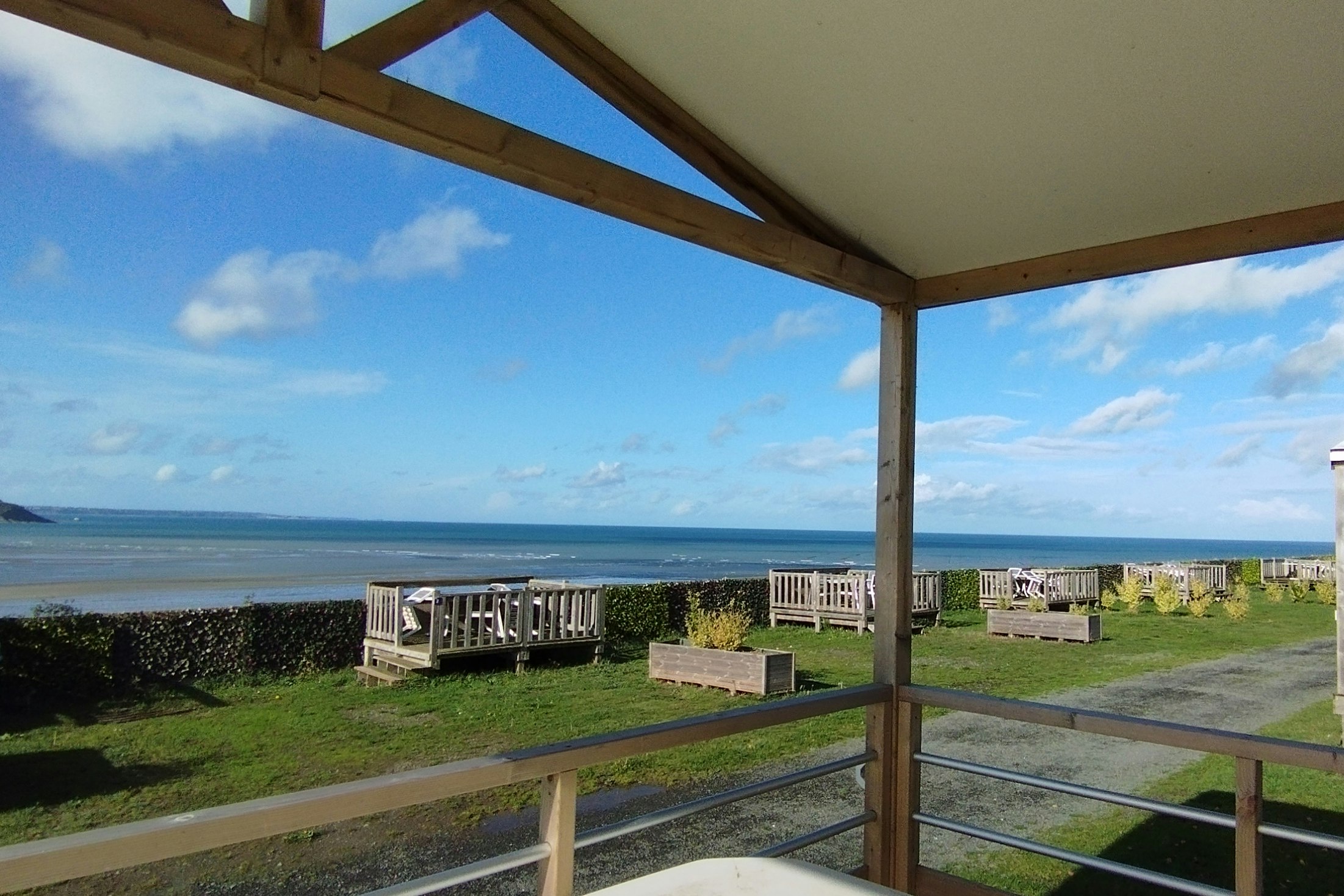 Blick von der überdachten Terrasse auf den Strand und das Meer, mit Sitzplätzen und kleinen Gartenwegen am Campingplatz.