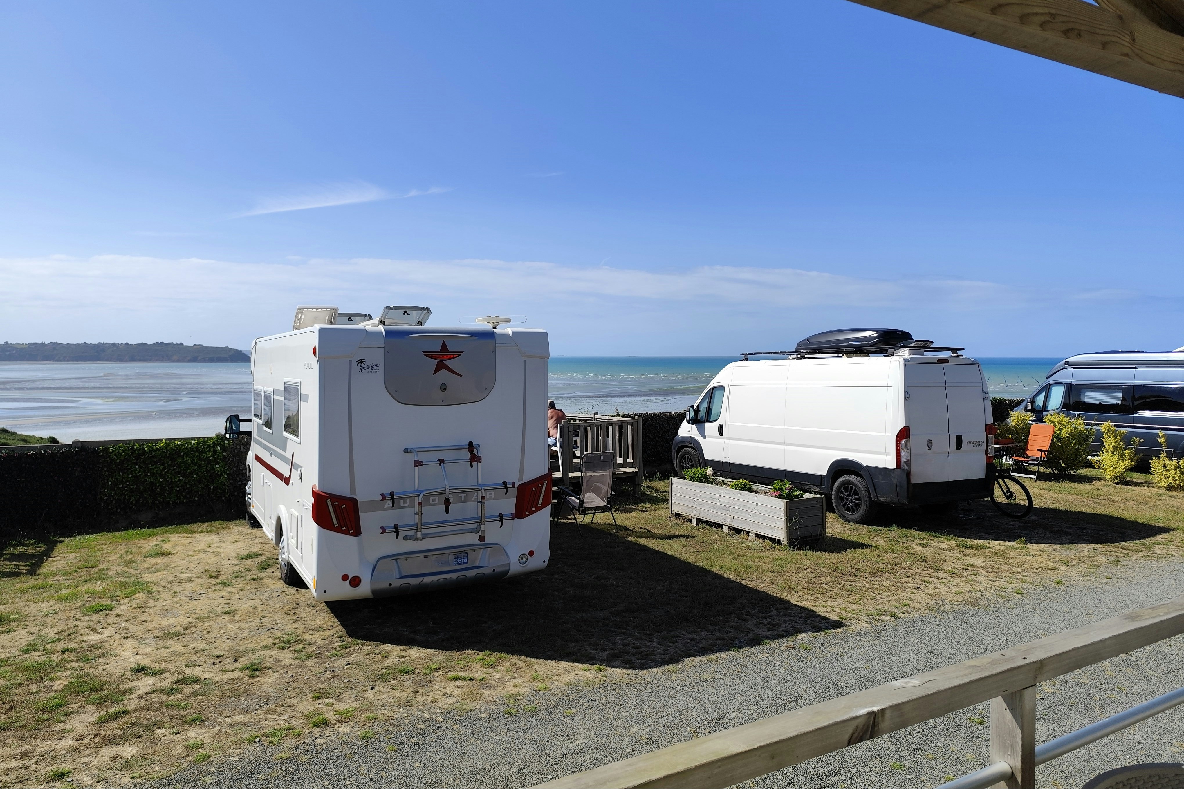 Wohnmobile auf einem Stellplatz am Meer, mit Blick auf Wasser und Küste, neben einem Holzgeländer.