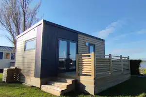 Moderner Camping-Bungalow mit Holzveranda, Sitzbank und kleinem Grill am Rasen, im Hintergrund Meer und Himmel.