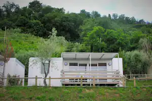 Campingunterkunft mit Terrasse aus Holz, weißen Wänden und großen Fenstern, umgeben von Bäumen und grünem Rasen.