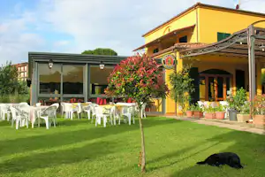 Gelbes Gebäude mit Terrasse, Markise und Garten mit Rasen, weißen Gartenstühlen, Tisch und einem schwarzen Hund.