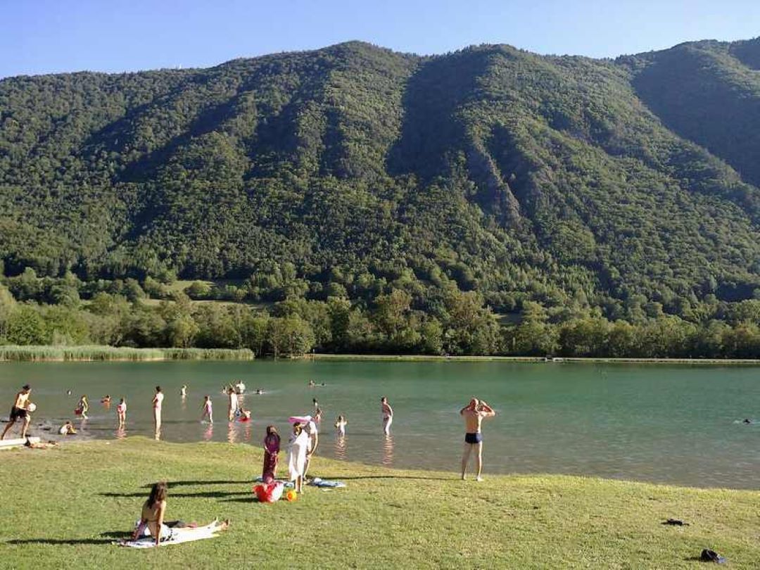 Menschen am Seeufer mit Liegestühlen und Handtüchern, im Hintergrund bewaldete Berge und grünes Ufer.