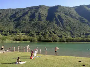 Menschen am Seeufer mit Liegestühlen und Handtüchern, im Hintergrund bewaldete Berge und grünes Ufer.
