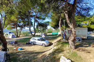 Campingplatz mit Wohnwagen, Zelt und Picknicktisch im Schatten der Bäume, daneben ein Auto auf dem Stellplatz.