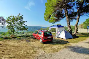 Zelt und rotes Auto auf einem Campingplatz zwischen Bäumen, mit Kiesweg und Rasenfläche im Hintergrund.