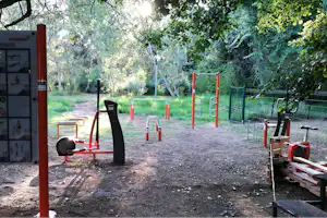 Spielplatz am Campingplatz mit orangefarbenen Fitness- und Klettergeräten, Sandboden und Zaun im grünen Wald.