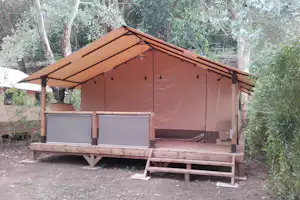 Zelt mit Vordach auf Holzpodest, seitliche Planen und Beleuchtung, umgeben von Bäumen und Sträuchern im Campingbereich.