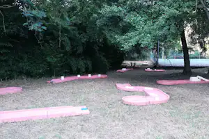 Minigolfanlage auf dem Rasen mit pinken Bahnen, Bäumen im Hintergrund und einem Zaun am Rand des Geländes.