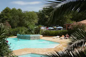 Campingpool mit türkisfarbenem Wasser, Palmen und Liegestühlen am Rand, im Hintergrund Bäume und Sonnenschirme.
