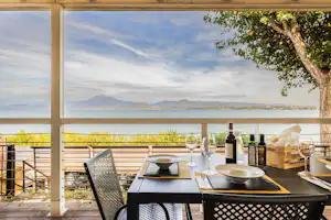 Terrasse am Campingplatz mit Esstisch, Stühlen und Flaschen, Blick auf den See und die Berge in der Ferne.