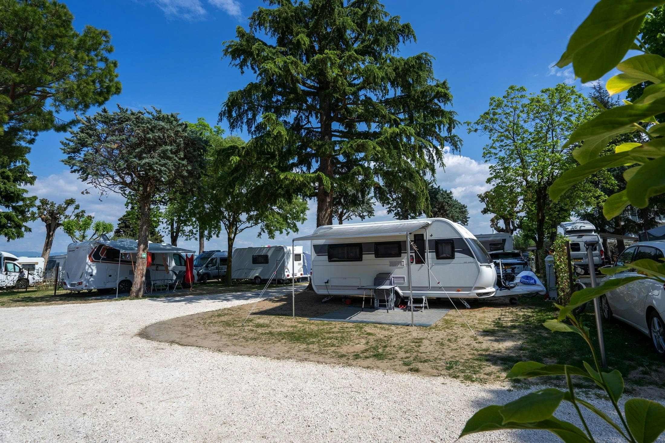 Wohnwagen auf einem Campingplatz zwischen Bäumen, mit Vorzelt und Sitzbereich, im Hintergrund weitere Wohnmobile.