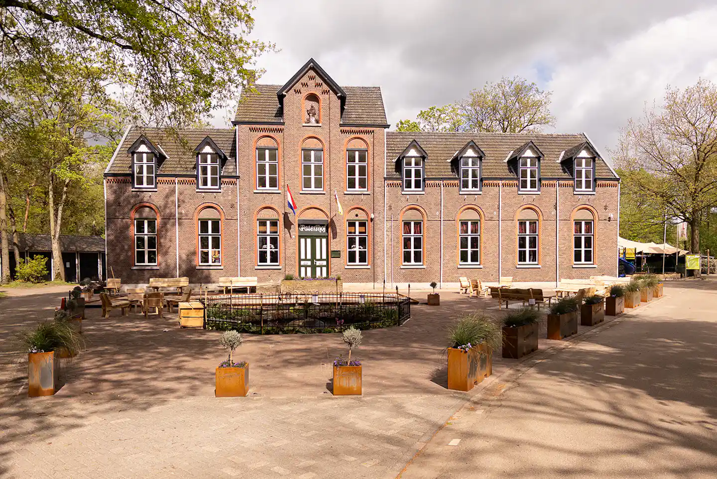Ziegelsteingebäude mit mehreren Fenstern und Terrasse, davor gepflasterter Platz mit Holzkisten und Bäumen am Rand.