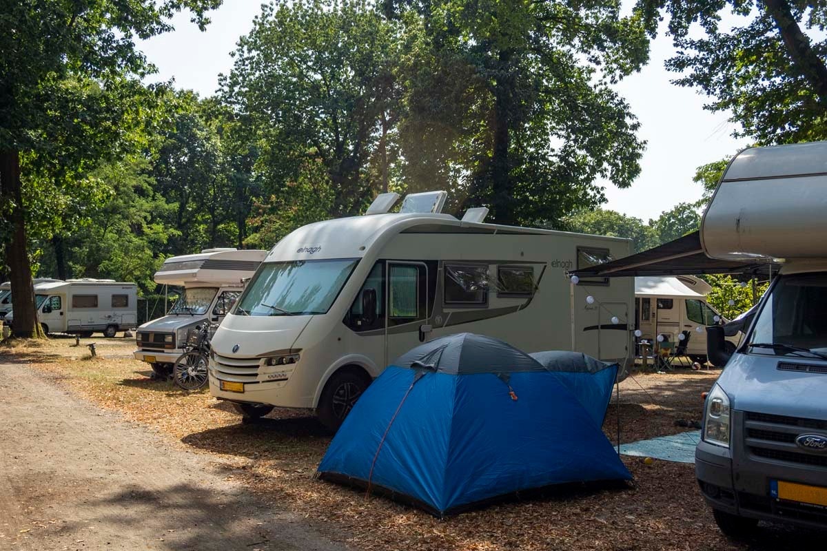 Blaues Zelt auf einem Campingplatz, daneben Wohnmobile und Wohnwagen, im Hintergrund Bäume und ein weiterer Stellplatz.