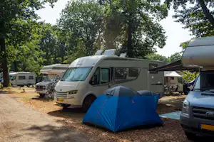 Blaues Zelt auf einem Campingplatz, daneben Wohnmobile und Wohnwagen, im Hintergrund Bäume und ein weiterer Stellplatz.