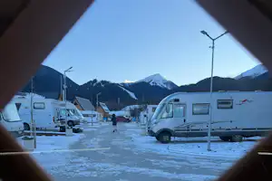 Wohnmobile und Caravans auf einem Campingplatz im Winter, Blick auf verschneite Berge und Laternen im Hintergrund.