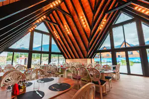 Innenraum eines Camping-Restaurants mit langen Holztischen, Stühlen und warmen Lichterketten, große Fenster mit Blick auf Berge und Wasser.