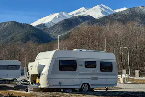 Weißer Wohnwagen auf einem Campingplatz mit Stellplatz, daneben ein weiterer Wohnwagen und Berge mit Schnee im Hintergrund.