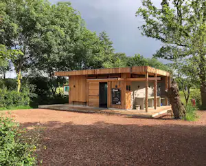 Holzhaus auf einem Campingplatz mit Terrasse, Holzfassade und Eingang, umgeben von Bäumen und Rasen.