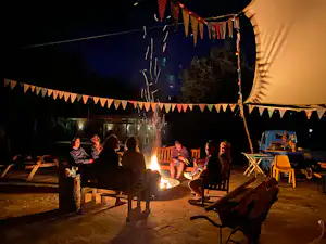 Zelt am Campingplatz mit Lichterkette, Lagerfeuer und mehreren Personen am Tisch und an Holzstühlen.