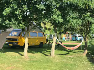 Gelber Camping-Transporter und Wohnwagen auf einem Stellplatz unter Bäumen, mit Hängematte und Rasen im Hintergrund.