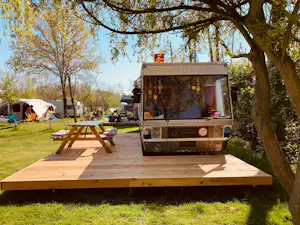 Wohnmobil auf einem Campingplatz mit Holzpodest, daneben Zelt und Picknicktisch im Grünen unter Bäumen.