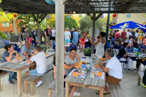 Camping-Restaurant unter einer Holzpergola mit langen Tischen, Speisen und vielen Gästen beim Buffet.