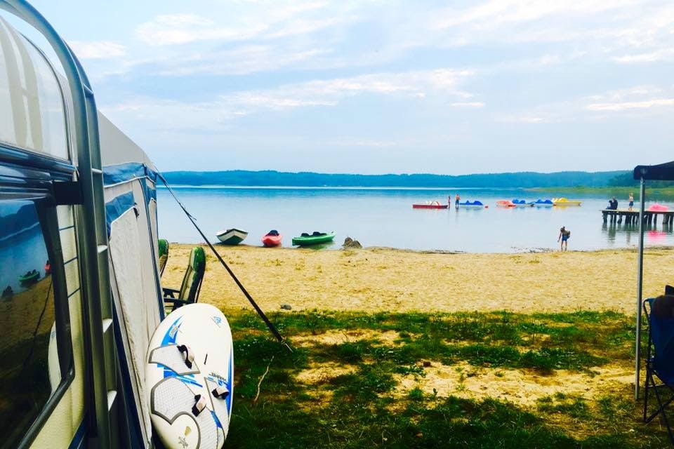 Zelt am Seeufer mit Surfbrett, Liegestühlen und Blick auf Paddelboote und Schwimmer im Wasser.