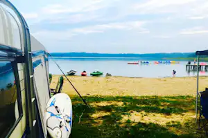 Zelt am Seeufer mit Surfbrett, Liegestühlen und Blick auf Paddelboote und Schwimmer im Wasser.