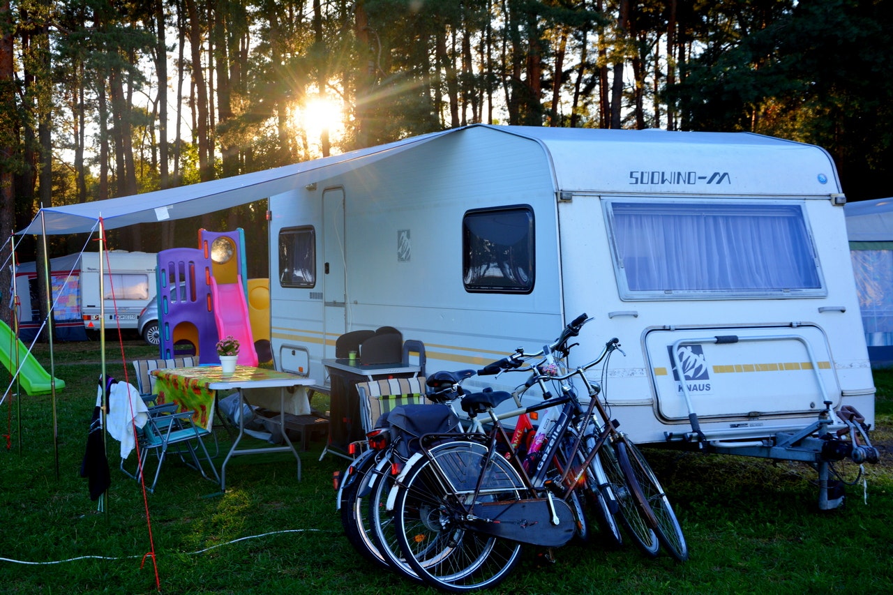 Weißer Wohnwagen im Campingwald, davor Fahrräder und Campingstühle unter einem Sonnenschirm, im Hintergrund Bäume und Abendsonne.