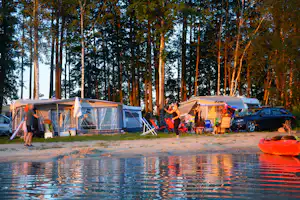 Campingplatz am See: Wohnwagen und Zelte mit Vorzelten, Sitzgruppe und Auto am Ufer zwischen Bäumen.