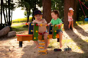 Kinder spielen auf einem Spielgerät im Sandbereich am Campingplatz, im Hintergrund Bäume und ein weiterer Spielbereich.