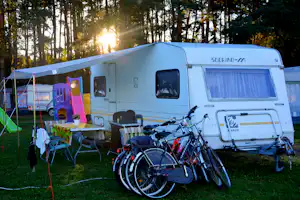 Weißer Wohnwagen mit Fahrrädern davor, Campingstühle und Tisch auf dem Stellplatz im Wald bei Sonnenuntergang.
