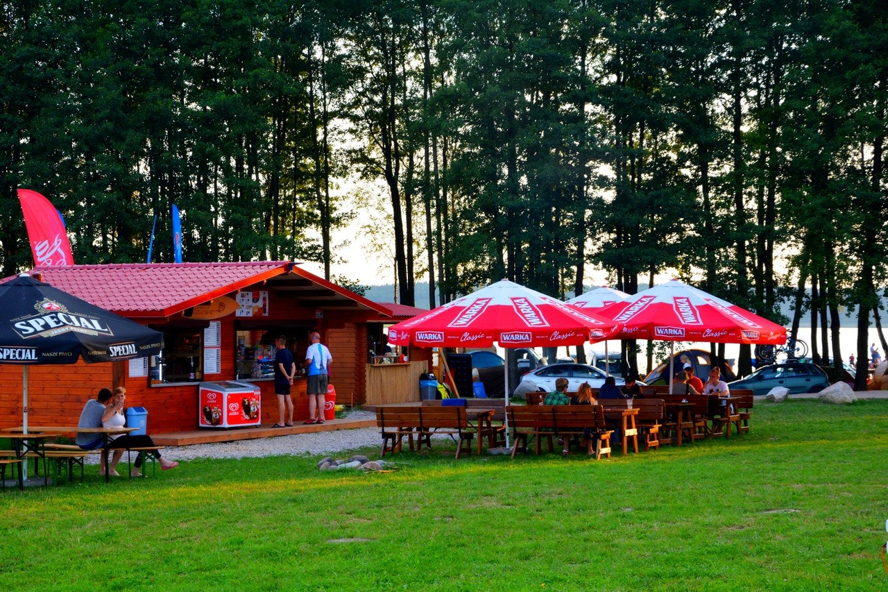 Terrasse am Campingplatz mit Holzhütte, roten Sonnenschirmen und vielen Tischen, umgeben von Bäumen im Grünen.