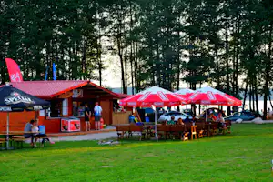 Terrasse am Campingplatz mit Holzhütte, roten Sonnenschirmen und vielen Tischen, umgeben von Bäumen im Grünen.