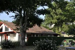 Campinghaus mit rotem Dach, großer Baum im Vordergrund und Zelt am Gartenbereich mit Stühlen und Blumen.