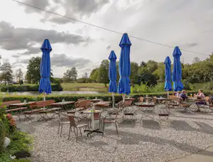 Terrasse mit vielen Metallstühlen und Tischen, blauen Sonnenschirmen und Blick auf Bäume und Wiese.