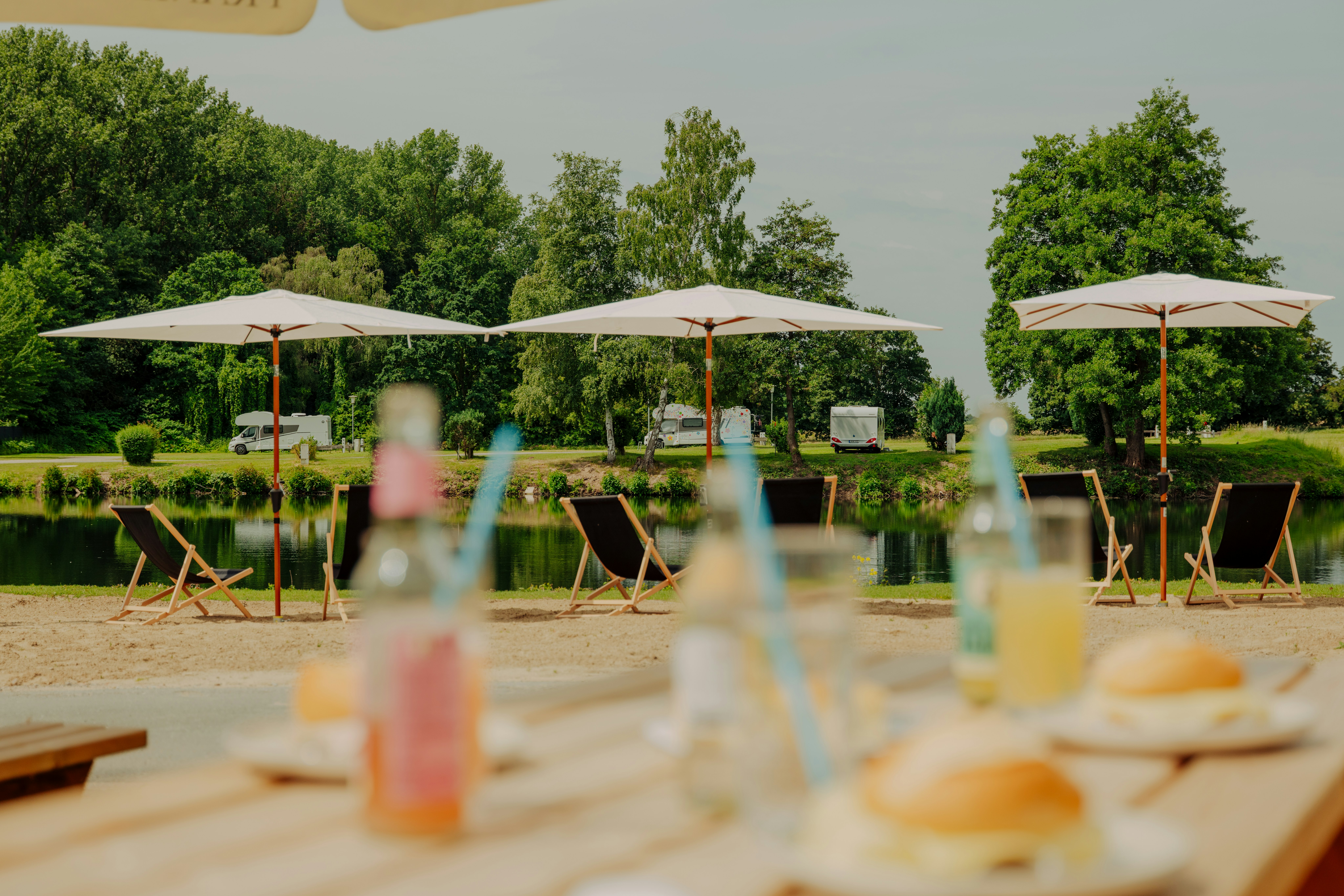 Tisch mit Getränken und Frühstück auf einer Terrasse, im Hintergrund Zelte, Liegestühle und Sonnenschirme am ruhigen Seeufer.