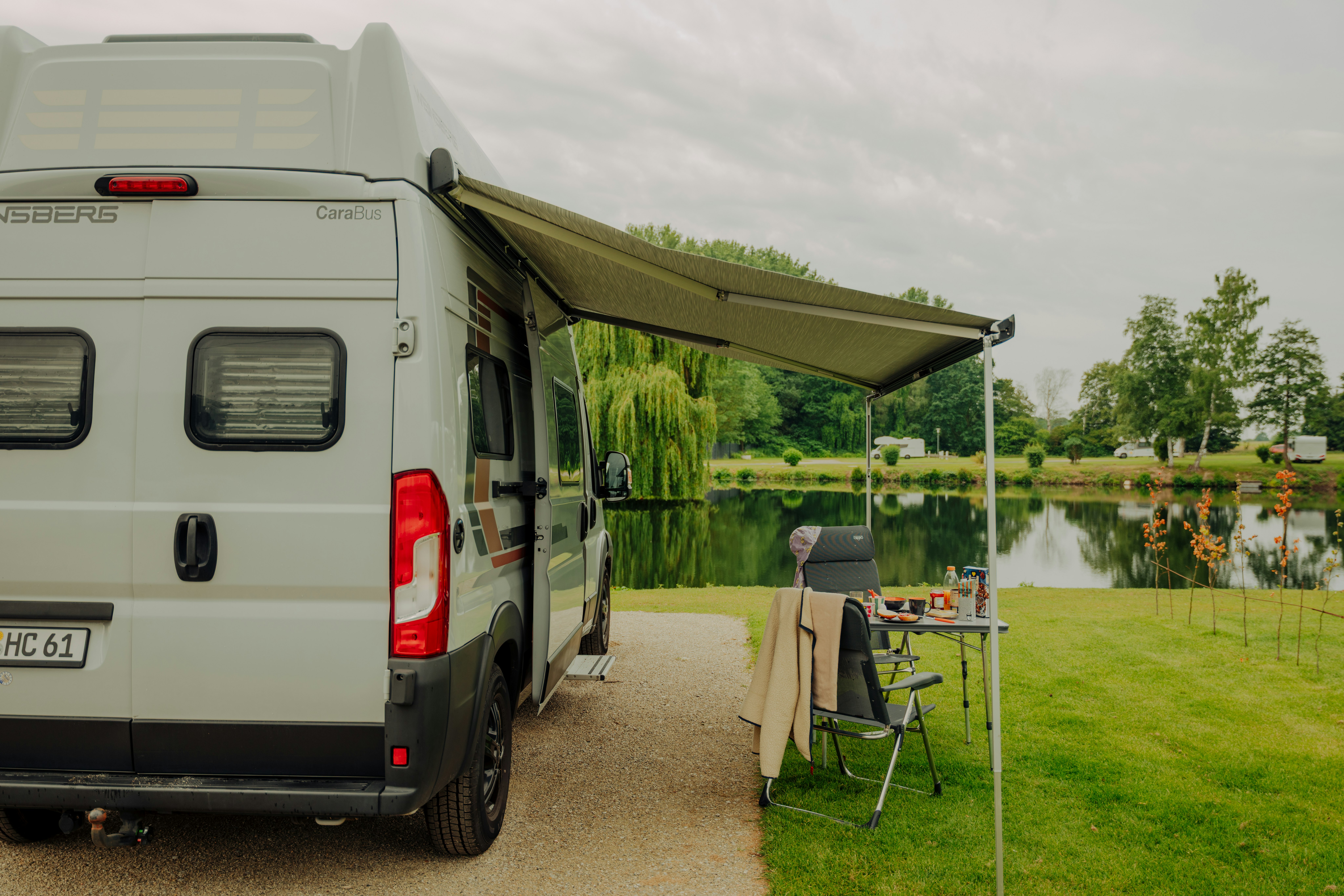 Wohnmobil am Stellplatz mit Markise, Campingstuhl und Tisch am Weg, im Hintergrund Rasen und See.