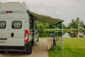 Wohnmobil am Stellplatz mit Markise, Campingstuhl und Tisch am Weg, im Hintergrund Rasen und See.