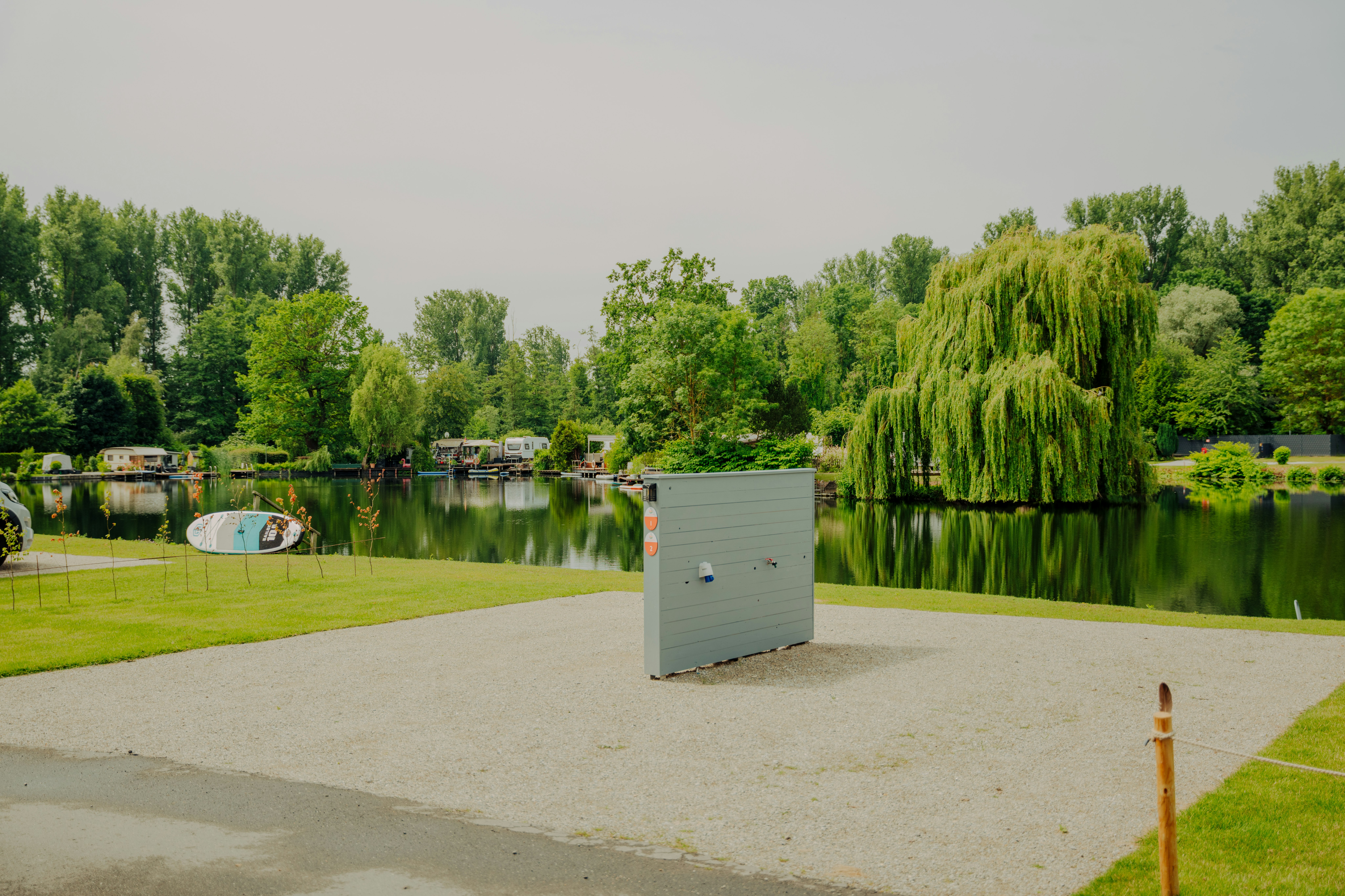 Ruhiger Blick auf einen Campingplatz am See mit Kiesweg, Rasenfläche, Bäumen und einem kleinen Gebäude im Vordergrund.