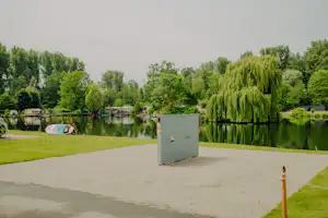 Ruhiger Blick auf einen Campingplatz am See mit Kiesweg, Rasenfläche, Bäumen und einem kleinen Gebäude im Vordergrund.
