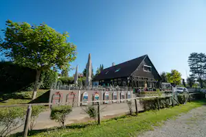 Campinghaus mit Holzfassade und Terrasse, davor Stühle und Sonnenschirme, umgeben von Bäumen und Hecken.