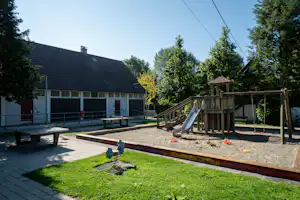 Spielplatz mit Rutsche, Sandfläche und Holzpodest, daneben Tische und Gebäude mit dunklem Dach, umgeben von Bäumen.