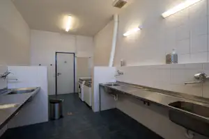 Waschraum mit Spülbecken, Armaturen, Waschmaschine und Abzugsschacht, gefliest mit hellen Wänden und Tür im Hintergrund.