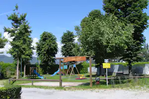 Spielplatz im Grünen mit Rutsche, Schaukel und Holzschild am Campingplatz, umgeben von Bäumen und Rasen.