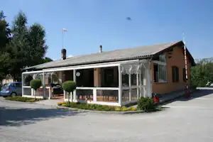 Wohnwagen mit verglaster Veranda und Sitzgruppe vor einem Campinghaus, umgeben von Büschen und Bäumen.